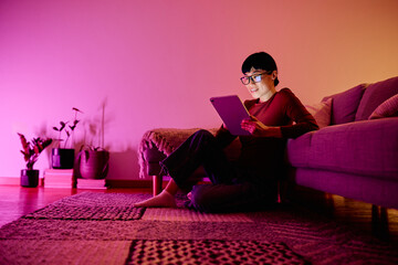 Young woman relaxing on the floor, holding tablet with glowing screen, enjoying content in cozy living room at night © we.bond.creations