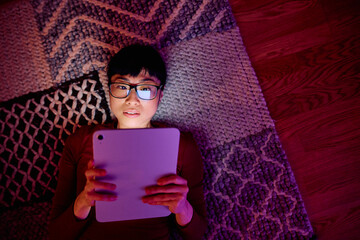 Young woman lying on rug at home using digital tablet in dark room, screen light illuminating face and glasses © we.bond.creations