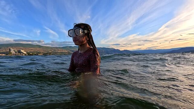 A young girl in long sleeve swimwear with leopard print pattern and snorkeling goggles standing and playing in water, slow motion.