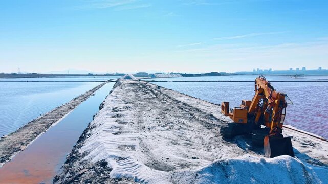 Yellow excavator on salt ridge between pink water ponds. Industrial scene with an excavator parked on a white salt path at a saltern with city skyline.
