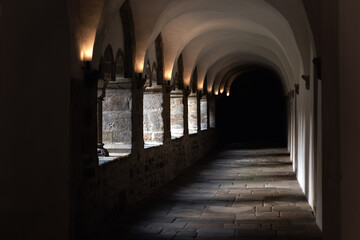 Fototapeta premium Medieval cloister corridor stone arches vaulted ceiling darkness illuminated warm sconces textured masonry wall. Perspective shadow silence create sacred atmosphere inside historic European monastery