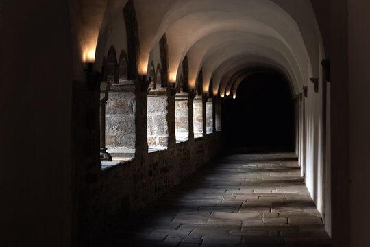 Medieval cloister corridor stone arches vaulted ceiling darkness illuminated warm sconces textured masonry wall. Perspective shadow silence create sacred atmosphere inside historic European monastery