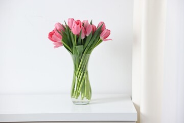 Fototapeta premium Bouquet of pink tulips in vase on chest of drawers near white wall indoors