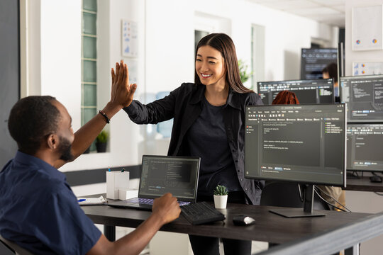 Colleagues celebrate coding project completion, cooperation and teamwork, capturing positive, professional workspace. African american and asian programmers smiling and gesturing in startup office.