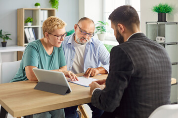 Senior couple sits with advisor reviewing and signing a contract and related document, formalizing...