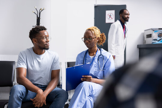 Young male adult sits with healthcare professional in modern medical facility, discussing recent medical test results. Nurse uses notes on clipboard to guide patient through health insurance details.