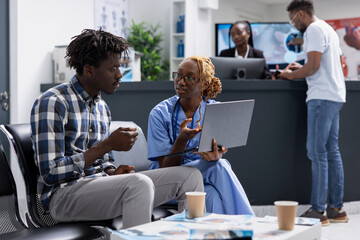 Obraz premium Black man patient sits with healthcare specialist in medical facility, engaged in focused conversation about test results. Nurse using laptop to assist clinic visitor with health insurance questions.