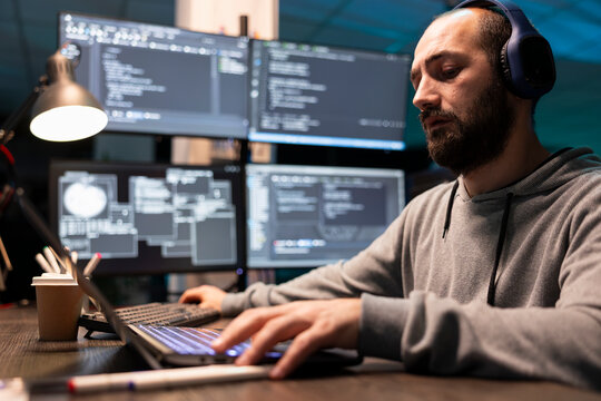 Bearded man wearing headphones concentrates on laptop while glowing monitors display technical interface. Freelance programmer listens to music and works on software development code in office.