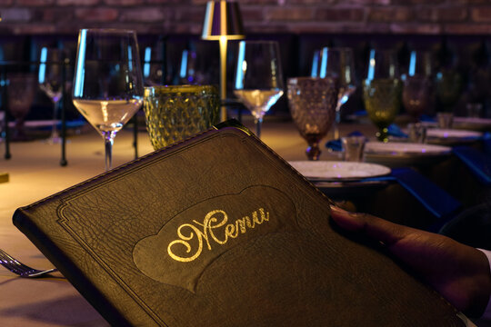 Closeup hand holding leather bound menu while seated at elegantly set restaurant table. Fine dining atmosphere with table setting, glassware, ambient lighting, concept of choosing food, hospitality