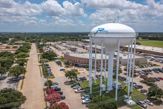 Aerial Drone View of Tall White Water Tower in Parking Lot With Parked Cars Along Street With Trees in Richardson, Texas