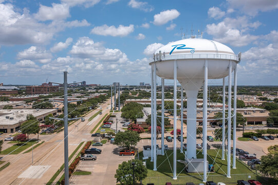 Aerial Drone View of Tall White Water Tower in Parking Lot With Parked Cars Along Street With Trees in Richardson, Texas