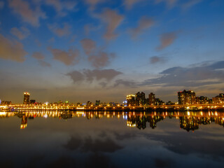 Naklejka premium City skyline lights reflect on calm river at twilight.