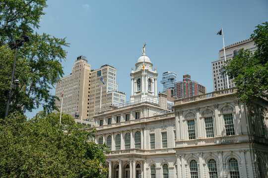 Detailed view of the historic New York City Hall with its iconic white clock tower. Surrounding trees and modern skyscrapers highlight the contrast of old and new architecture.