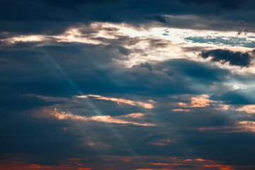 Dramatic crepuscular rays piercing through dark, layered stratocumulus clouds at sunset. Sun is setting, casting a warm glow on the clouds in the sky