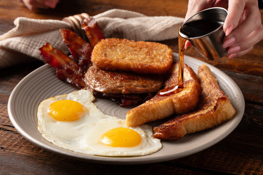 Close-up rustic shot of a woman pouring syrup on French toast on a plate with eggs, bacon, sausage, and hash browns. 