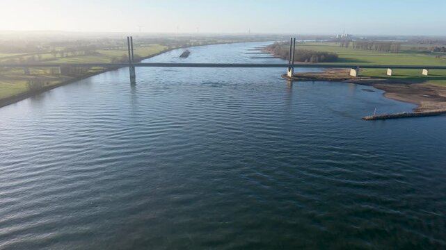 Aerial view of a wide river crossed by a modern bridge, with a barge moving along calm water, surrounded by green fields, sparse trees, and distant wind turbines in a rural landscape.