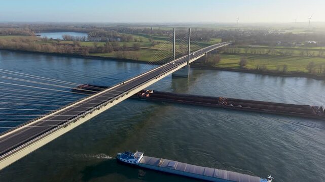 Aerial view of a cable stayed bridge spanning a wide river as two large vessels pass below, surrounded by green fields, forest patches, and distant wind turbines across a calm rural landscape.