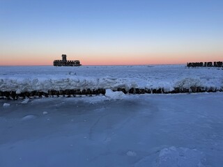 Snow‑covered wooden breakwater and an abandoned structure standing on a frozen sea during sunrise. Soft pink and blue tones in the sky create a calm winter atmosphere.