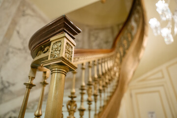 A close-up of an ornate golden stair railing features a floral carving and polished wooden handrail. The elegant curved staircase in the background glows under a crystal chandelier.