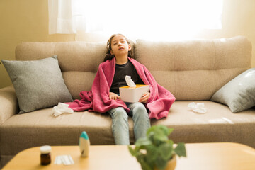 Obraz premium Tween girl feeling sick at home with flu symptoms, covered in blanket and holding tissues