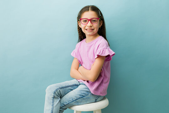 Young tweenage girl with pink glasses and braids smiling confidently, posing for a studio portrait on a vibrant background