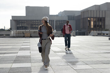 Diverse students walking across plaza, female wearing headphones holding phone and rolled posters