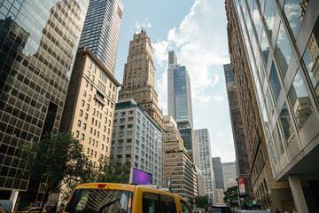 A yellow taxi drives down a bustling street in Midtown Manhattan. Historic and modern skyscrapers rise into a bright summer sky.