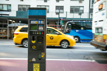 A New York City parking meter stands in focus as a yellow taxi, bus, and car blur past in the background. The image captures urban life and motion in Manhattan.