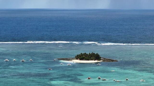 Island with palm trees and sandy beach surrounded by shallow coral reef and boats. Guyam Island. Siargao, Philippines.