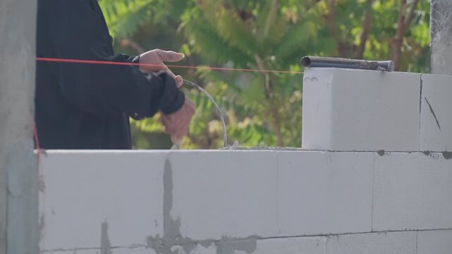 Close up of construction worker applying cement mortar on concrete block wall using string line for precision alignment. Professional masonry process at outdoor building site.