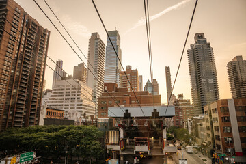 The Roosevelt Island Tramway glides between tall skyscrapers in Midtown Manhattan during golden hour. The sky is warm and glowing, highlighting the city's iconic skyline.