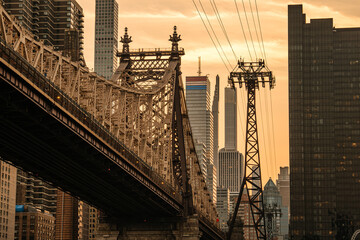 A detailed view of the Queensboro Bridge structure at sunset with Manhattan skyscrapers rising in the background. The golden sky enhances the contrast between historic architecture and modern high