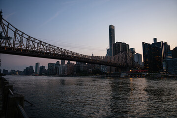 The Queensboro Bridge stretches across the East River as the last light of day fades over Manhattan's skyline. Reflections shimmer on the calm water, and the city prepares for nightfall.