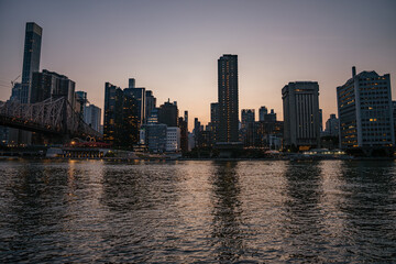 The Manhattan skyline is seen across the East River during golden hour, with high-rise buildings glowing in the fading light. The water reflects the warm tones of the sunset and the city's illuminated