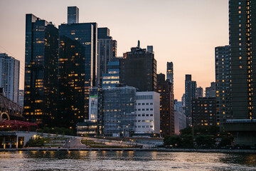 A close view of modern glass skyscrapers and older concrete buildings in Manhattan during sunset. The glowing reflections and city lights create a vibrant evening cityscape.