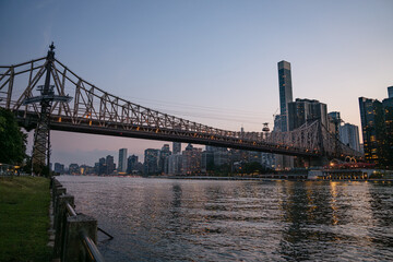 The Queensboro Bridge stretches across the East River as the Manhattan skyline lights up in the evening. Viewed from the riverside path, the scene captures the calm water and towering buildings under