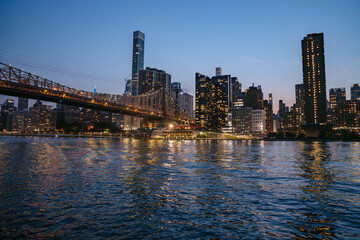The Queensboro Bridge and Manhattan skyscrapers glow with golden lights at night, reflected in the calm East River. The peaceful scene captures the contrast of sparkling architecture and dark water.