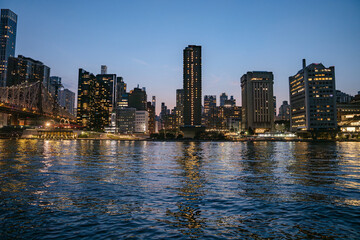 The Manhattan skyline glows against a deep blue sky as city lights shimmer on the surface of the East River. Tall towers and modern buildings line the waterfront, creating a vibrant evening scene.