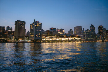 Modern office and residential buildings along the Manhattan waterfront glow brightly against the night sky. Their lights shimmer across the surface of the East River, creating a vibrant city