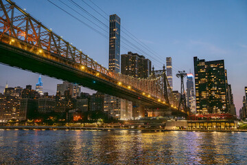 The Queensboro Bridge stretches across the East River, illuminated by warm lights during twilight. Modern skyscrapers of Midtown Manhattan glow against the evening sky, reflecting beautifully on the