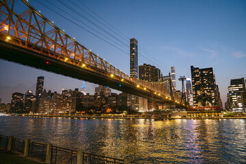 The Queensboro Bridge glows with lights as it spans across the East River, framing the New York City skyline at twilight. Skyscrapers and reflections create a striking urban evening scene viewed from