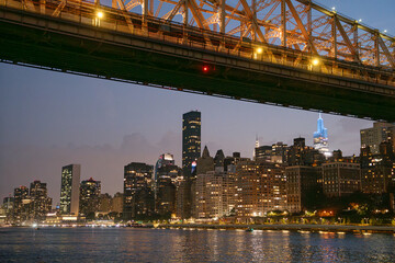 The Manhattan skyline glows at dusk beneath the illuminated structure of the Queensboro Bridge. Skyscrapers and waterfront lights reflect on the calm East River, creating a classic New York evening