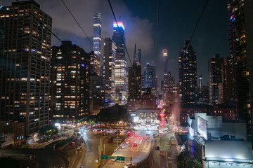 The Roosevelt Island Tram glides above a glowing Midtown Manhattan street filled with traffic and skyscrapers. Bright lights, illuminated buildings, and a dramatic skyline define this vibrant New York