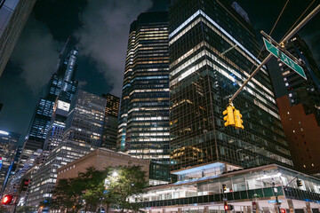 Tall glass skyscrapers tower above East 53rd Street in New York City, illuminated by office lights and traffic signals. A yellow streetlight and dark sky add contrast to the vibrant urban night scene.