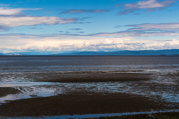 Obraz premium Low‑tide sandy beach with puddles reflecting a partly cloudy sky and distant mountains on Vancouver Island, BC.