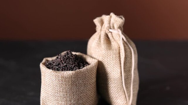 Woman pouring dried black tea leaves in burlap sack at table, closeup