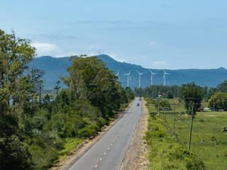 Obraz premium Rural road leading to wind turbines with mountain landscape under blue sky