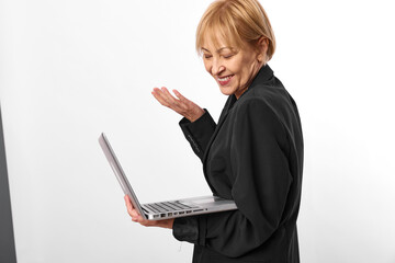 Smiling mature woman in black jacket works on laptop, looking at screen with positive expression. Concept of remote work, business, technology, and female professional in studio.