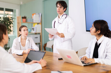Obraz premium Confident young guy medical student standing near table where sitting interns talking and discussing ideas during meeting in bright office