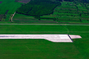 Aerial shot captures concrete runway. Airfield is surrounded by expansive green fields and dense patches of forest, characteristic of its rural landscape